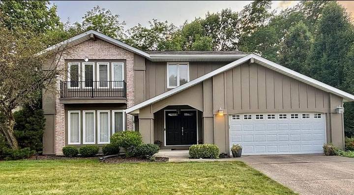 Original daytime listing photo of the front of the house: taupe siding, brick wing, balcony, black double doors and a white two-car garage