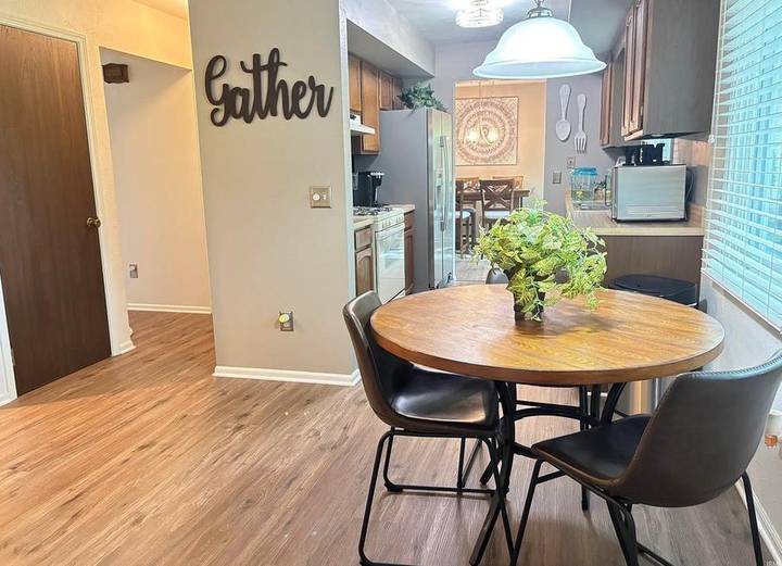 Original listing photo of the breakfast nook: round wood table, a Gather sign on the wall, and the galley kitchen beyond