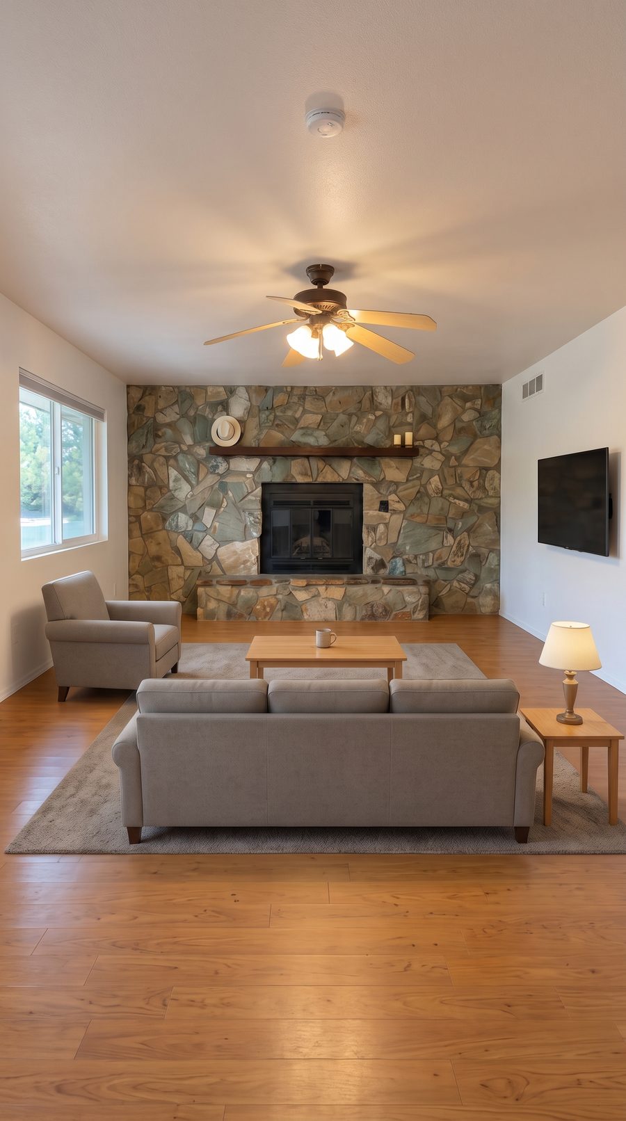 The same living room virtually staged: the same stone fireplace, fan, floor, and wall TV, now with a neutral sofa, armchair, coffee table, rug, and lamp